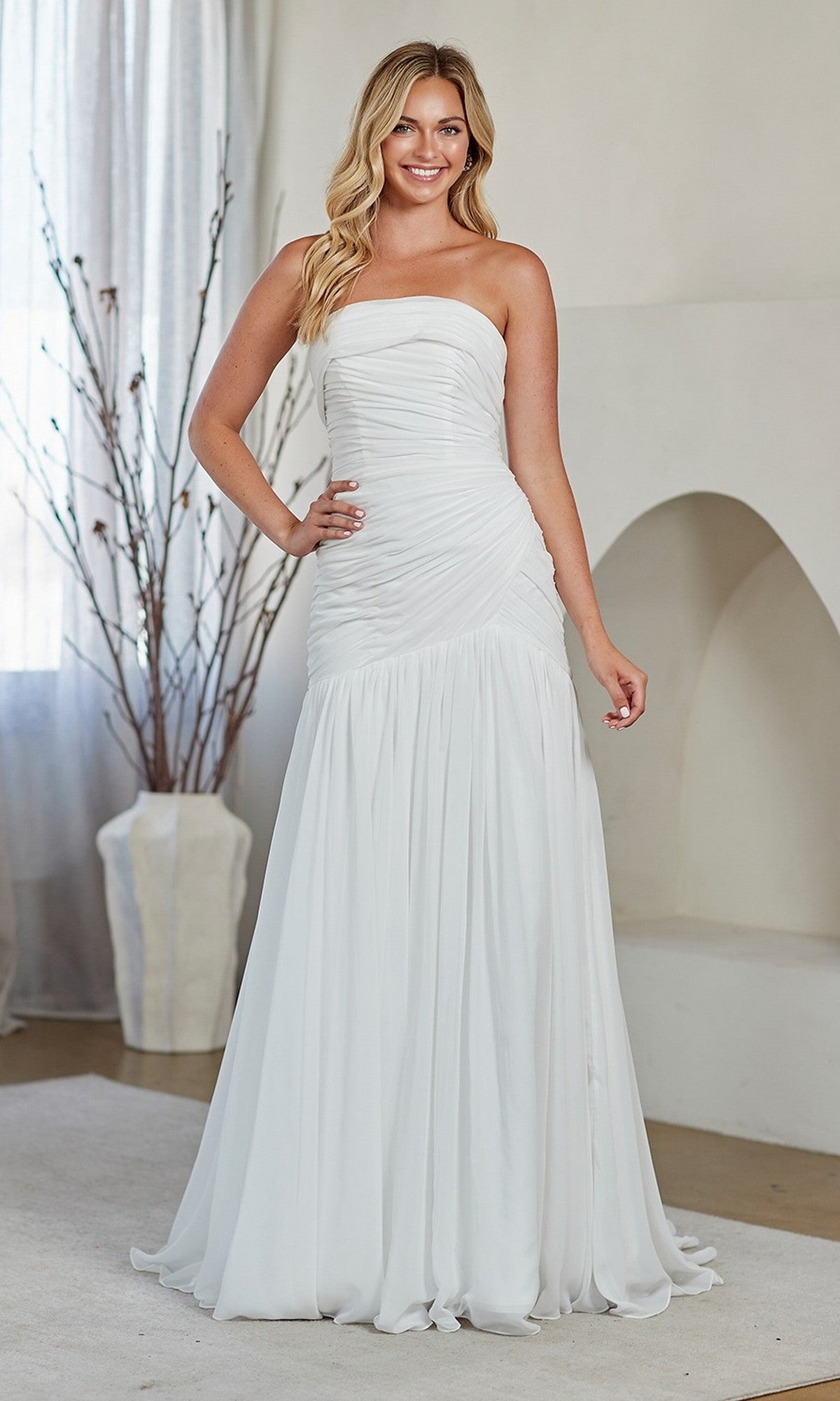 A woman stands indoors, smiling in a white Amelia Couture 6132 long prom dress featuring a strapless style and figure-flattering ruching on the bodice.
